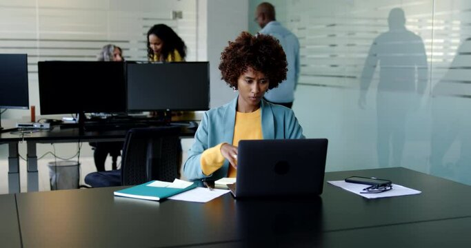 Young businesswoman writing notes and working on a laptop at her desk in an office with female colleagues talking in the background