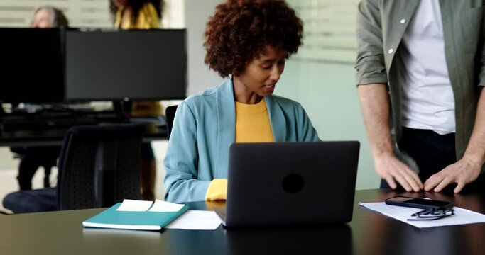 Young businesswoman talking with a male colleague while writing notes and working on a laptop at her office desk
