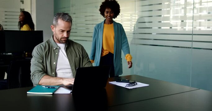 Businessman talking with a female colleague while working on a laptop at his desk in an office