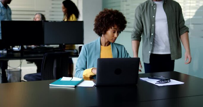 Young businesswoman talking with a male coworkers while writing notes and using a laptop at her desk in an office