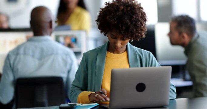 Young businesswoman writing notes while working at her desk in an office with colleagues talking in the background