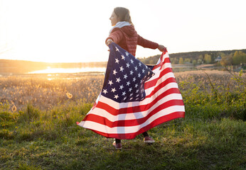 happy girl carelessly stands at sunset in the sun and holds the US flag behind her back....