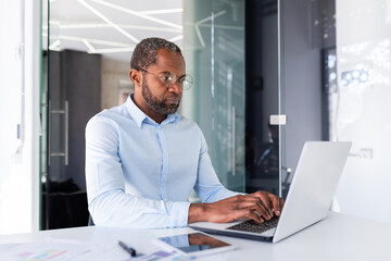 Serious and focused african american businessman in shirt working inside office with laptop, mature...