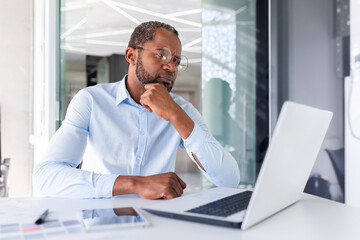 Thinking serious African American businessman working inside office, successful business man boss looking at laptop screen pondering important technical decision.