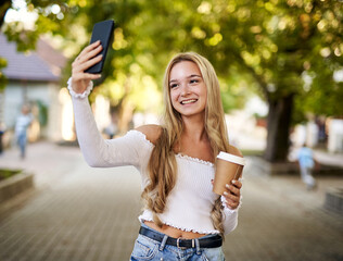 Young teeanager girl walking on a town street and taking selfie while holding takeaway paper coffee...