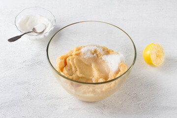 A glass bowl of pureed quince, lemon juice and sugar, half a lemon and a bowl of sugar on a light gray background. Stage of preparation of vegan quince pastille or other dessert.