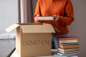 charity, donation and volunteering concept - young woman puts books in cardboard box at home - obrazy, fototapety, plakaty