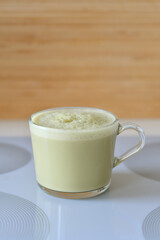 Matcha latte with oatmilk in a glass mug or cup with a neutral and blurry background. Matcha latte is yellow or green in color and has foamy and frothy oatmilk.