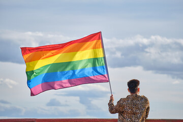 non-binary young asian person from behind holding a rainbow gay pride flag blowing in the wind