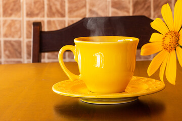 Uma xícara amarela com café quente, saindo vapor, com um girassol, sobre uma mesa de madeira.