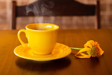 Uma xícara amarela, com café quente saindo vapor, com um girassol murcho sobre uma mesa de madeira e encosto da cadeira desfocada ao fundo.