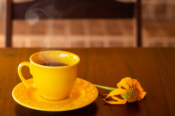 Uma xícara amarela, com café quente saindo vapor, com um girassol murcho sobre uma mesa de madeira e encosto da cadeira desfocada ao fundo.