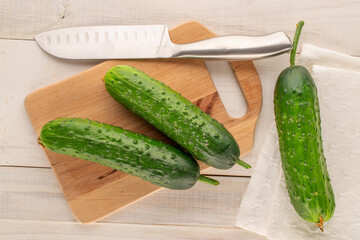 Three ripe green cucumbers with wooden kitchen board and metal knife on wooden table, macro, top view.