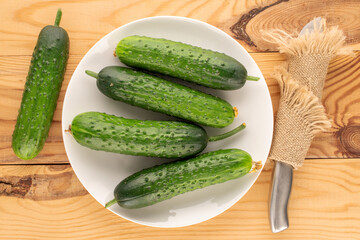 Five ripe green cucumbers with white ceramic plate and knife on wooden table, macro, top view.