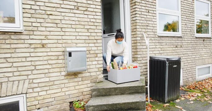 Woman in a protective face mask bringing a grocery delivery box into her home from her doorstep
