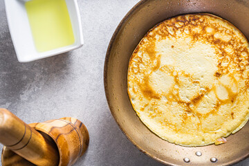 Delicious pancakes in a frying pan on a white table, with wheat, eggs, olive oil, tomato sauce and a checkered tablecloth in the background.