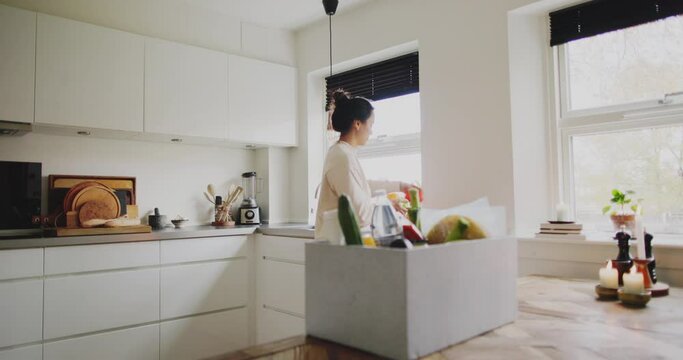 Young woman unpacking a delivery box full of groceries while standing at a table in her kitchen