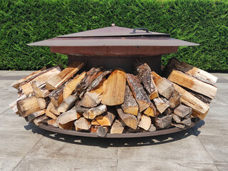 Round metal fire bowl. Rusty old iron brazier for making grill with stacked wood on the lower tier. Outside wood fireplace with a cover against the backdrop of green bushes. Firewood for a barbecue