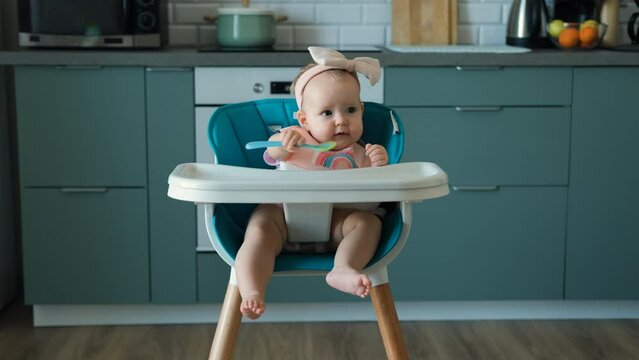 Cute baby girl waiting for food in high chair in the kitchen, playing with silicone spoon  Generative AI