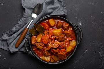 Beef meat stew with potatoes, carrot and delicious gravy in black casserole pot with bay leaves with spoon on black dark rustic concrete background from above . Generative AI - obrazy, fototapety, plakaty