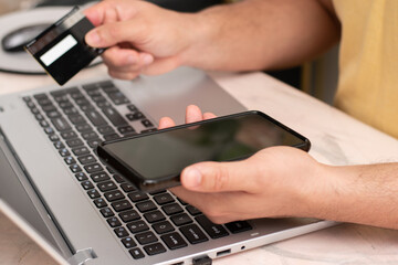 Young man buying with a black credit card and his phone front the laptop, online pay and shopping.