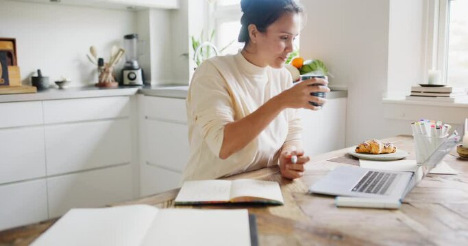 Young woman drinking tea while drawing in a sketchbook and working on a laptop at her kitchen table