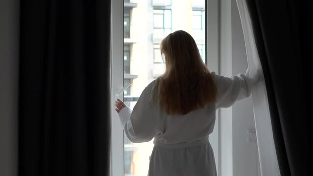 A beautiful and young girl in a white coat comes to the window and opens the curtains (curtain)