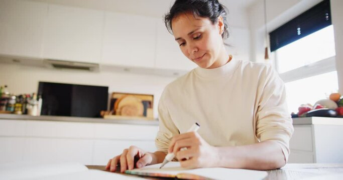 Smiling young woman sketching designs in a book while sitting at a table in her kitchen at home
