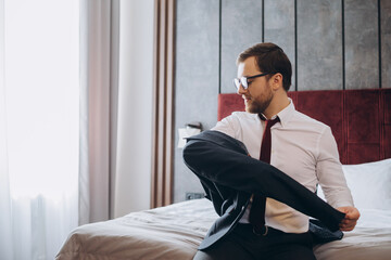 Business man with suitcase taking off jacket while sitting on bed in hotel room