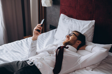 A businessman rests lying on a bed in a hotel room. A young man uses his smartphone while resting.