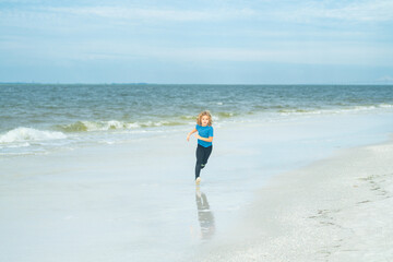 Cute happy little kid running along the sea coast outdoor on summer beach. Little kid run on summer beach. Excited child run against blue sky and sea. Summer vacation concept. Kids play at the sea.