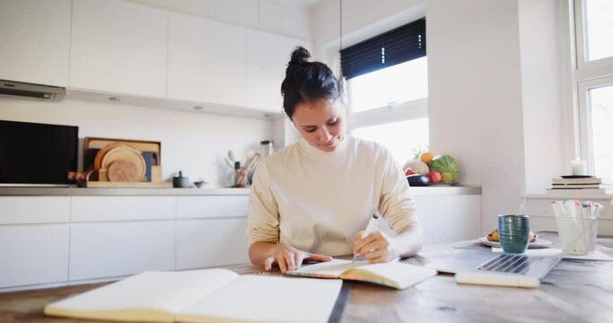 Young woman smiling while sitting at a table in her kitchen at home and sketching designs in a book
