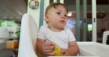 Thoughtful pensive baby infant on highchair holding healthy corn snack