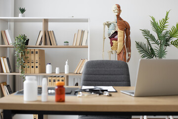Focus on human anatomy model on stand near office shelving with books and pills bottles at consulting room with writing desk on blurred foreground. Comfortable professional workplace in health center.