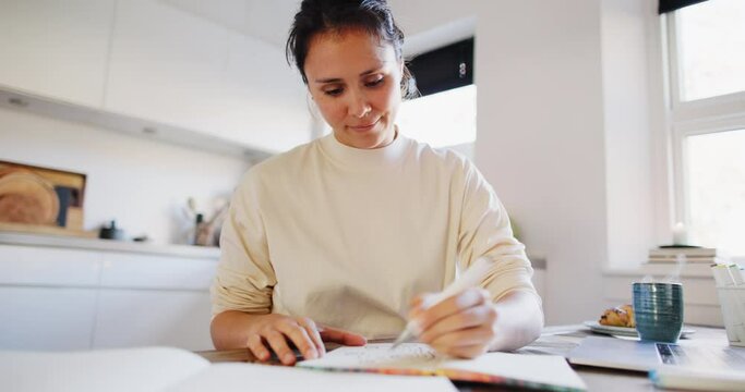 Smiling young woman working on some new design ideas in a sketchbook at a table in her kitchen at home