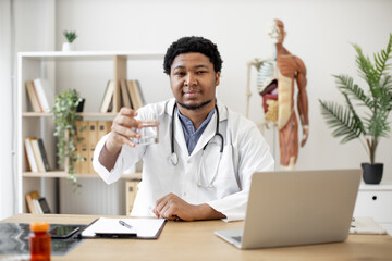 Obraz premium Positive multicultural physician in lab coat showing glass of water while sitting at writing desk with laptop and documents in doctor's office. General practitioner recommending staying hydrated.