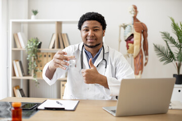 Confident multiethnic therapist at office desk holding half full glass and showing thumb-up sign while working in consulting room. Family doctor approving benefits of clean water for healthcare.