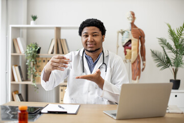 Positive multicultural physician in lab coat showing glass of water while sitting at writing desk...