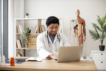 Attractive african american male in lab coat working on laptop while sitting at office desk in consulting room of medical clinic. General practitioner researching illnesses online in cozy workplace.