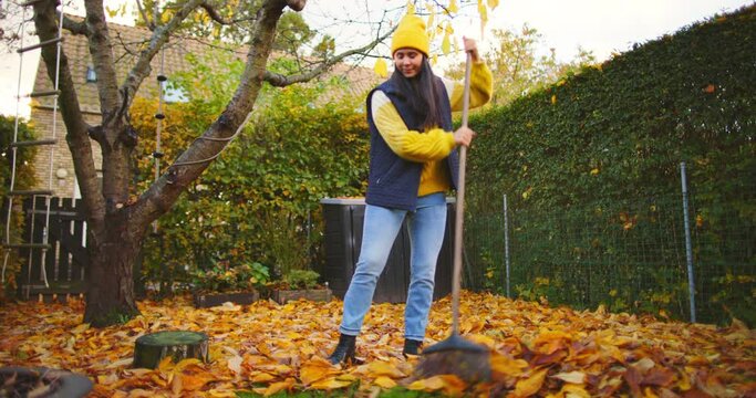 Smiling young woman raking up colorful leaves while cleaning up her backyard on a cool autumn afternoon