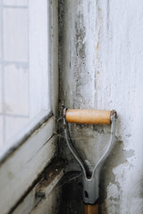 An abandoned, old shovel, garden tool with a wooden handle stands in the corner of the wall of the house.
