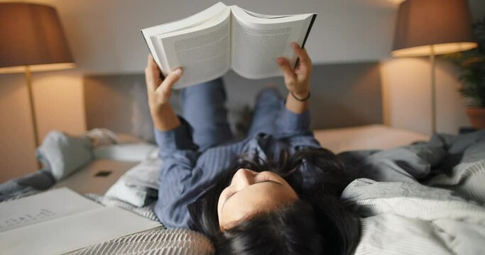 Young woman wearing pajamas reading a book while relaxing on her bed in the evening