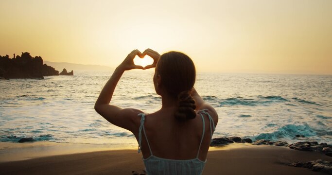 Woman on beach showing heart or love symbol with hands at golden sunset. Summer vacation travelers dream. Valentine's day background.