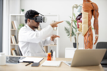 Side view of multiethnic male in white coat using virtual reality headset while sitting at office...
