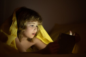 Kid is addicted to tablet, kid use tablet, watching cartoon. Child watching his tablet in the bed. Illuminated child face from device screen. Boy under the covers hold a tablet. Night time.