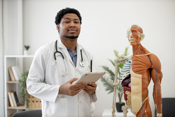 Portrait of positive african american man in white coat holding digital tablet while posing near...