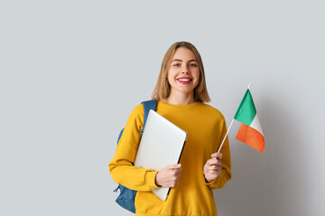 Young woman with flag of Ireland, laptop and backpack on grey background