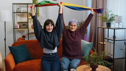 Two lesbian women family couple or girl friends holding LGBT people gay pride flag in hands at home...