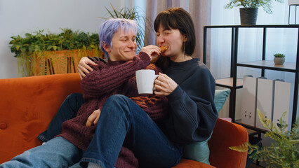Two lesbian women family couple, girl friends drinking coffee or tea, eating croissants and talking at home morning living room. LGBT same-sex people. Enjoying happy breakfast together lying on sofa