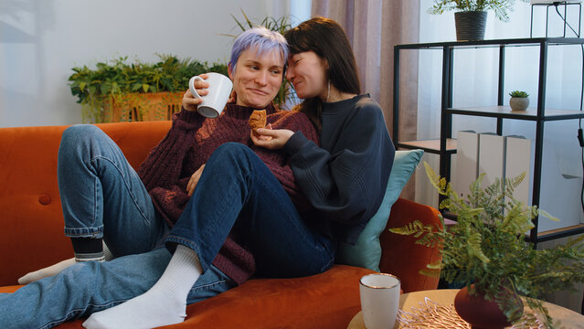 Two lesbian women family couple, girl friends drinking coffee or tea, eating croissants and talking at home morning living room. LGBT same-sex people. Enjoying happy breakfast together lying on sofa - Powered by Adobe